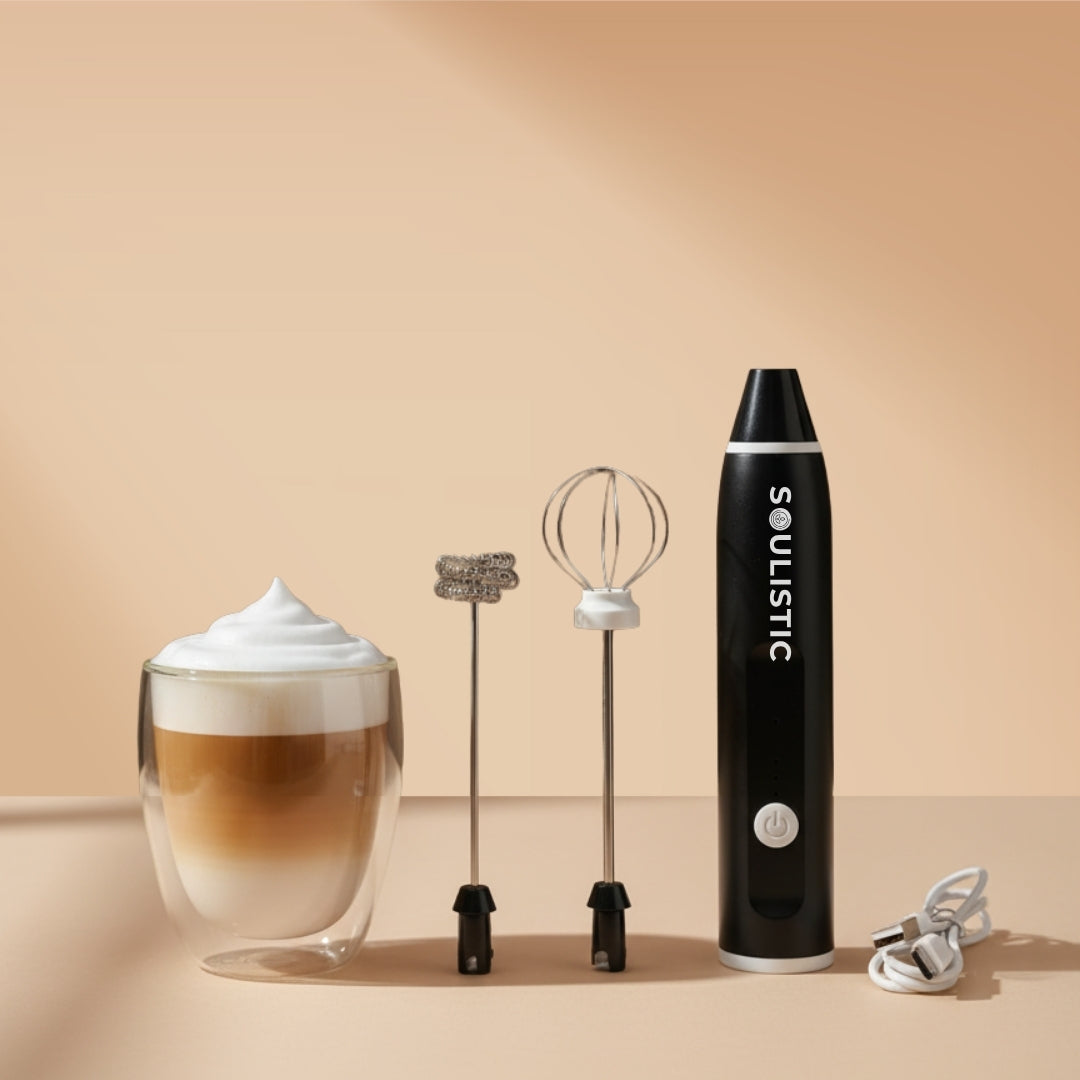 Soulistic Electric milk frother with its frothing and whisking attachments and USB to Type-C charging cord next to a double-walled glass of latte on a beige background.