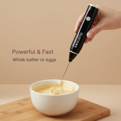 Hand using a black handheld Soulistic whisk to mix batter in a white bowl on a beige background